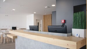 A reception desk with a wooden counter and two HP computers. There are chairs and tables in the background, and a plant is visible on the right. - HMB Fermentelos Hotels (Agueda)