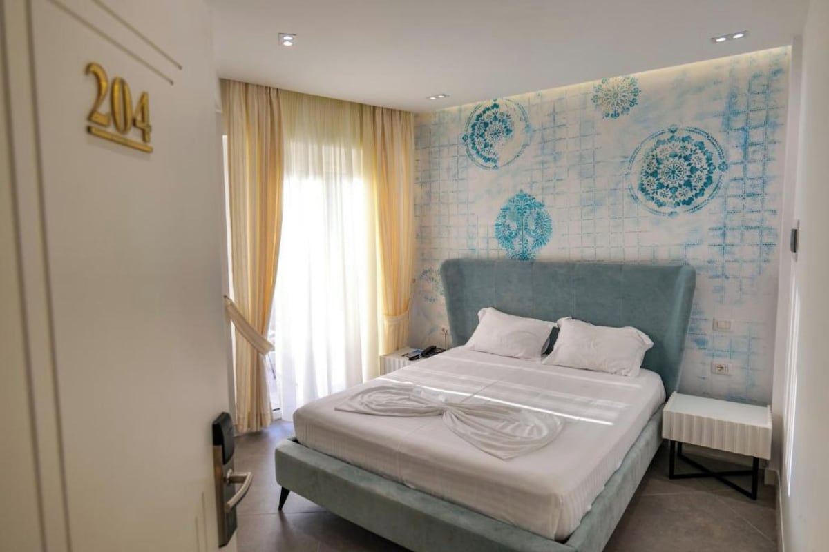 A hotel room with a bed, a bedside table, a wall with a decorative pattern, and a number sign on the wall.