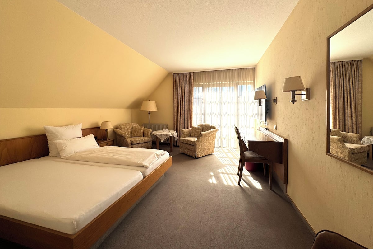 A hotel room with a large bed, two armchairs, a desk, and a mirror.