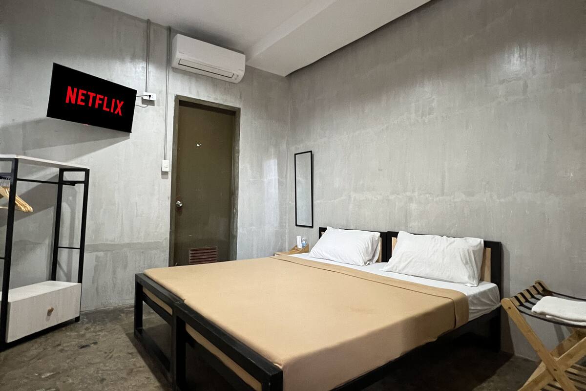 A bedroom with a bed, a bedside table, a chair, a wall-mounted TV, and an air conditioning unit.