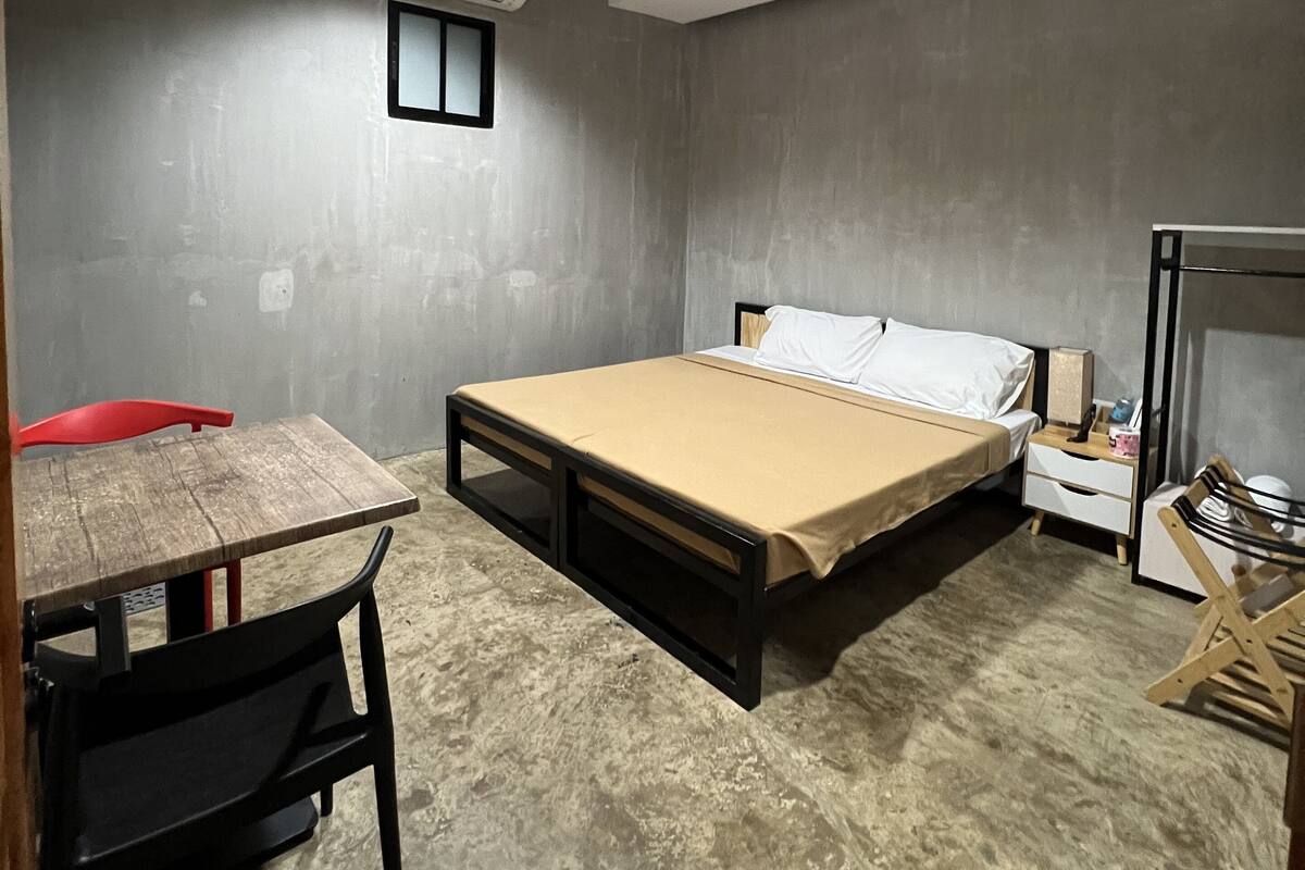A compact room with a bed, a small table, a chair, a nightstand, and an air conditioning unit.