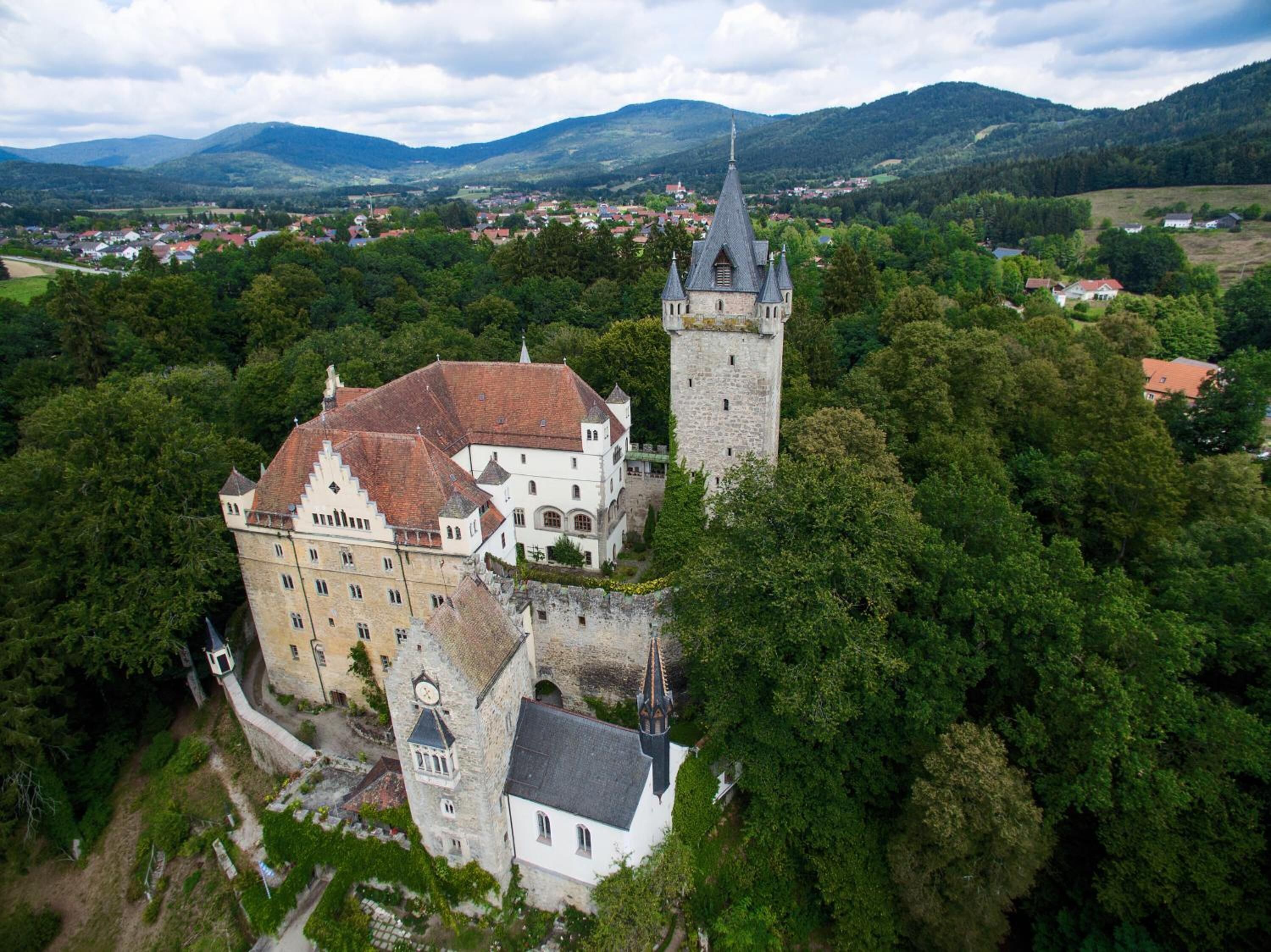 Vista aérea de um castelo histórico com uma torre imponente, rodeado por vegetação exuberante e um cenário montanhoso.