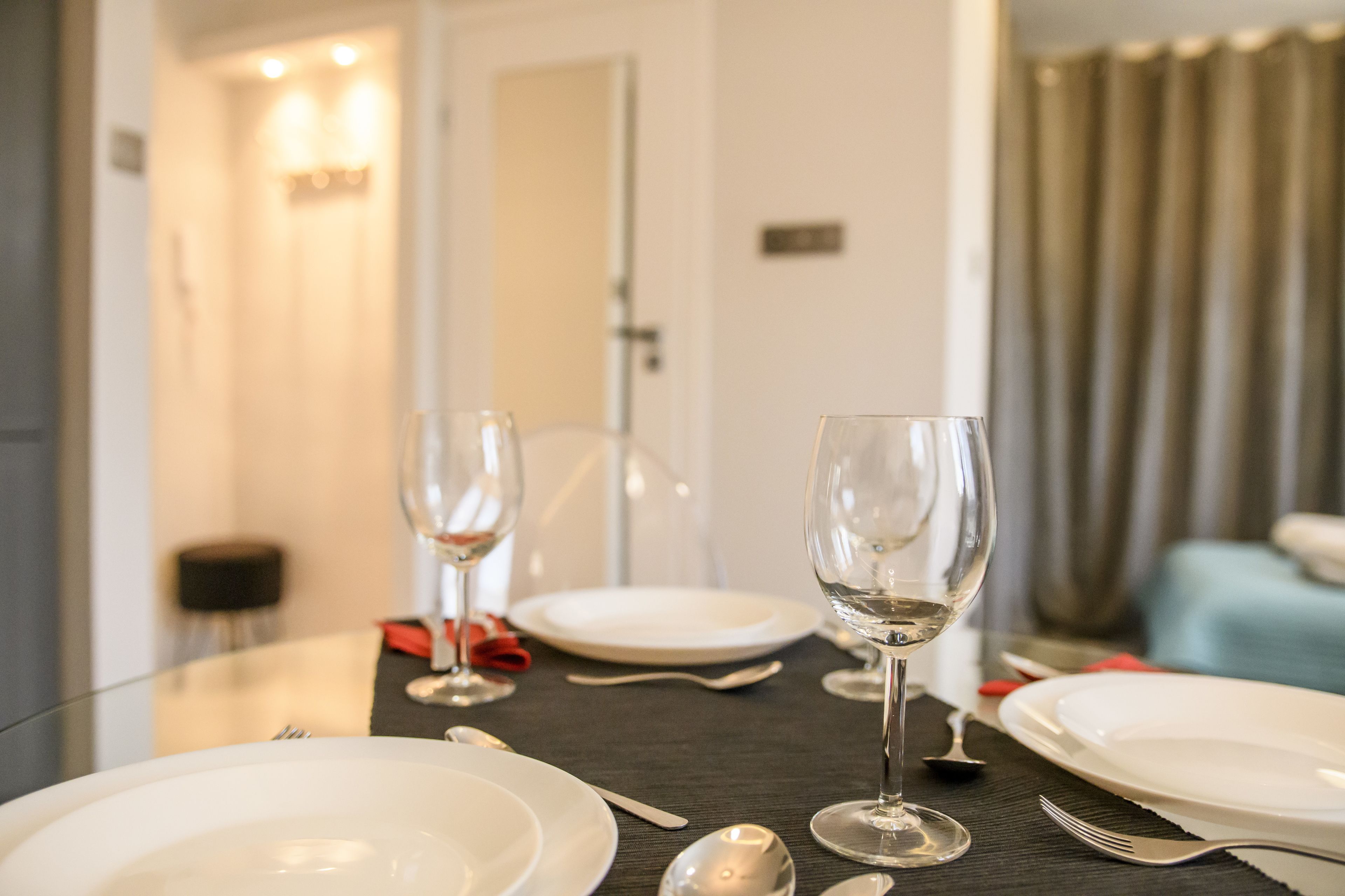 A dining table set with white plates, silverware, and wine glasses, with a view of a door and curtains in the background.