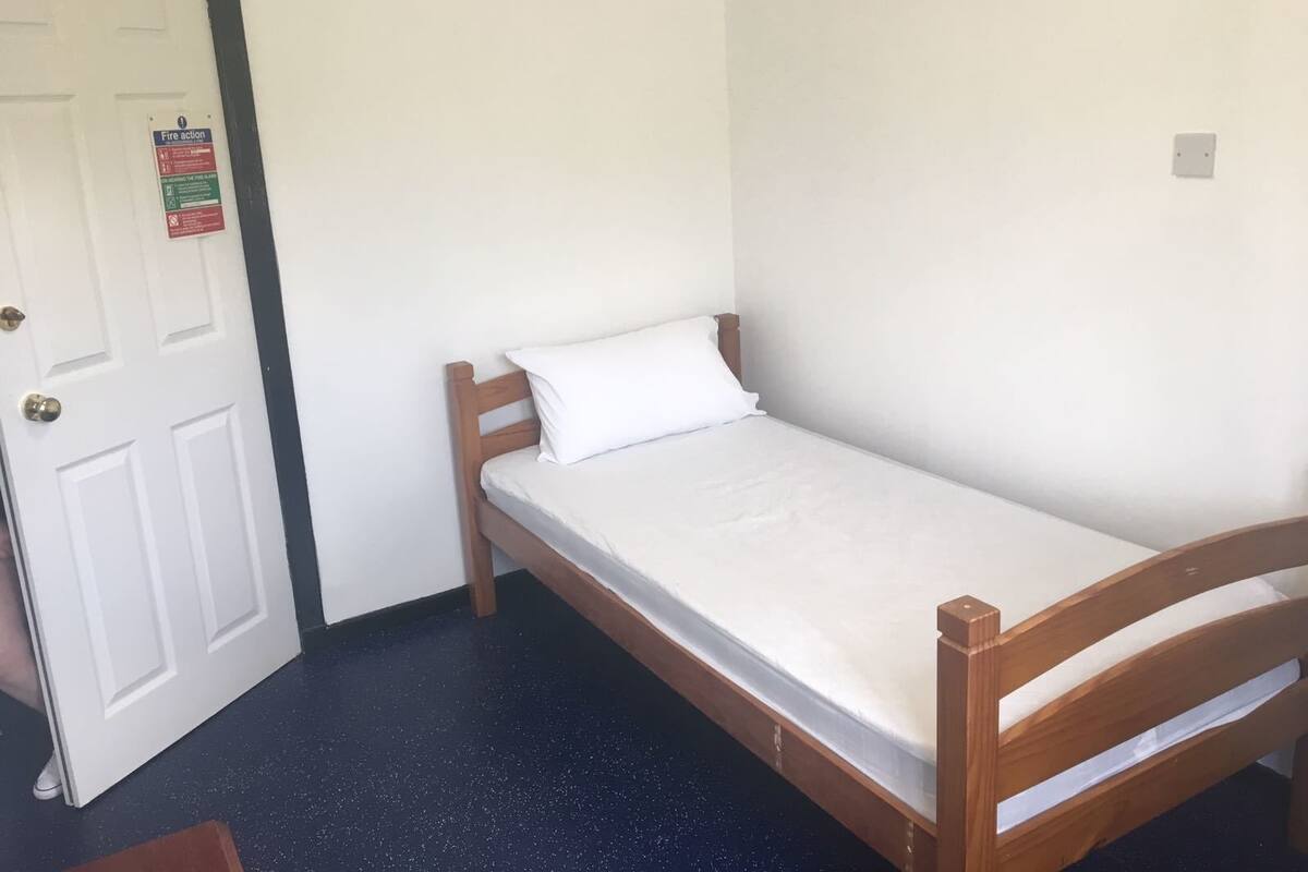 A single-occupancy room with a wooden bed, white bedding, a door with a fire action sign, and a dark speckled floor.