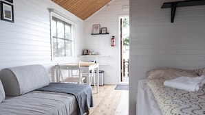 Una habitación acogedora con techo de madera, cama con cubrecama estampado, sofá con manta gris, una pequeña mesa con sillas y ventana con persianas.