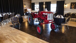Un restaurante con un tractor rojo como pieza central, paredes de madera y cortinas negras. Las mesas están cubiertas con manteles blancos y sillas doradas.