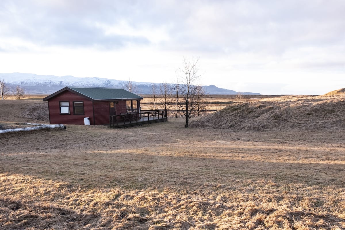 Une petite cabane rouge avec un toit vert, une terrasse en bois et une vue sur des montagnes enneigées au loin.