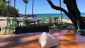 A beachside restaurant with outdoor seating, palm trees, and a view of the ocean.