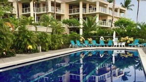 A swimming pool with lounge chairs, surrounded by palm trees and a multi-story building with balconies.