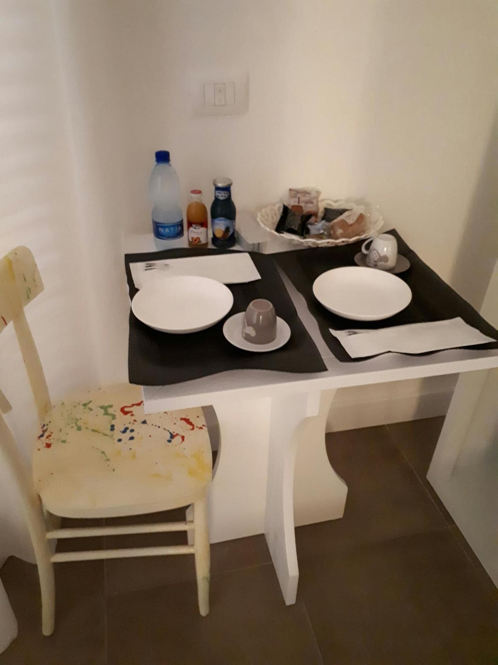 Un petit coin repas avec une table blanche dressée pour deux personnes, une chaise et un panier de pain.