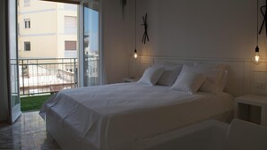Slaapkamer met een groot bed, nachtkastje, hangende plant en uitzicht op een gebouw via een balkondeur.
