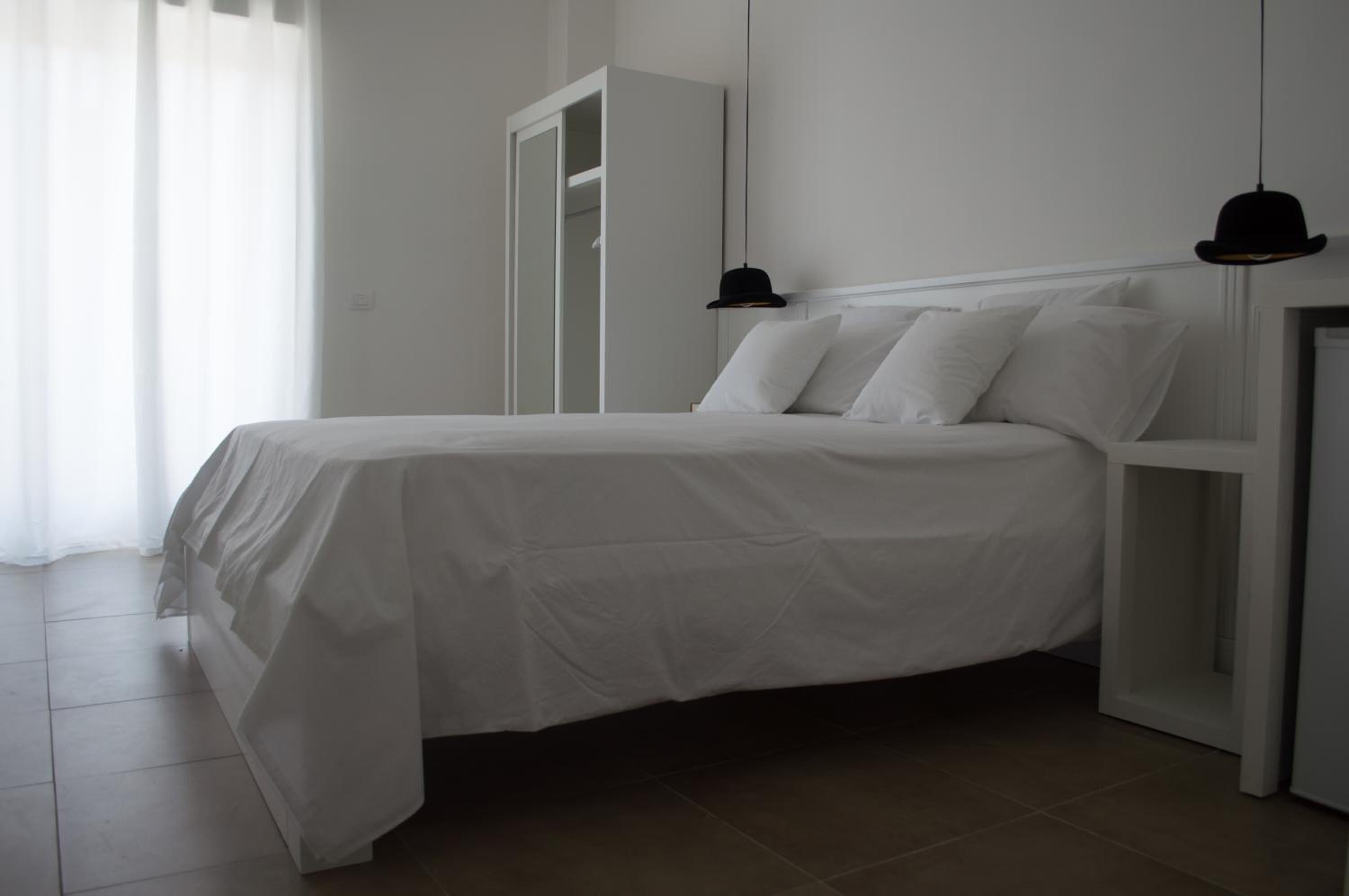 A neatly made bed with white linens in a room with a white wardrobe and a black hat hanging on the wall.