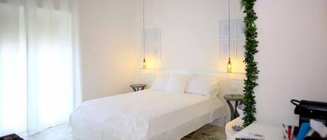 A hotel room with a bed, a small table, a chair, a desk, and a plant.