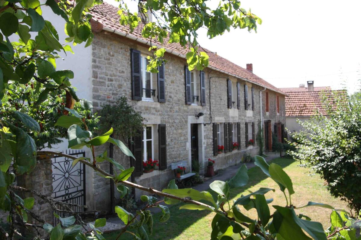 A stone building with multiple windows and shutters, surrounded by greenery and a small garden.
