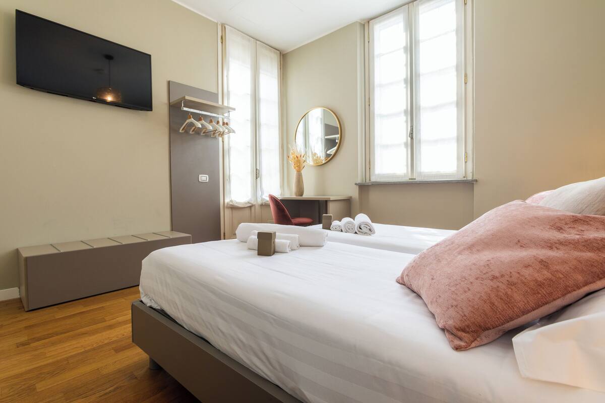 A modern hotel room with a large bed, a flat-screen TV mounted on the wall, a bedside table with a vase, and a window with curtains.