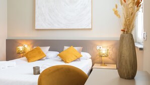 A neatly made bed with white linens and two mustard-colored pillows, flanked by two bedside lamps on white nightstands. A large abstract painting is hung above the bed, and a tall vase with dried plants is placed on a table beside the bed. - Martini Suite (Milan)
