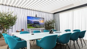 A conference room with a large screen, teal chairs, a white table, and a potted plant. - Martini Suite (Milan)