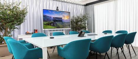 A conference room with a large screen, teal chairs, a white table, and a potted plant.