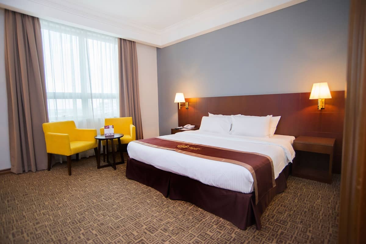 A hotel room with a large bed, two bedside lamps, a small table, a chair, and a window with curtains.