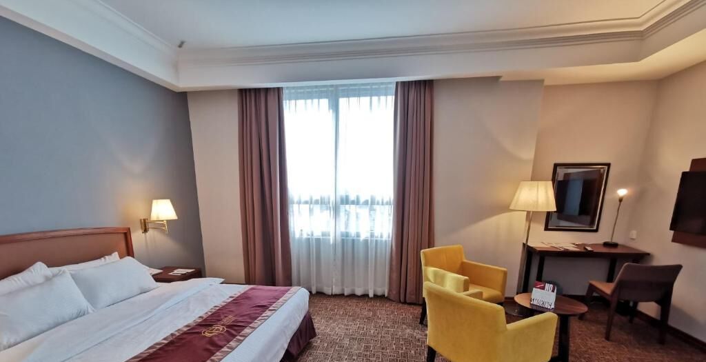 A hotel room with a bed, two chairs, a desk, a TV, and a window with curtains.