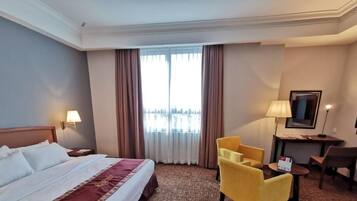 A hotel room with a bed, two chairs, a desk, a TV, and a window with curtains.