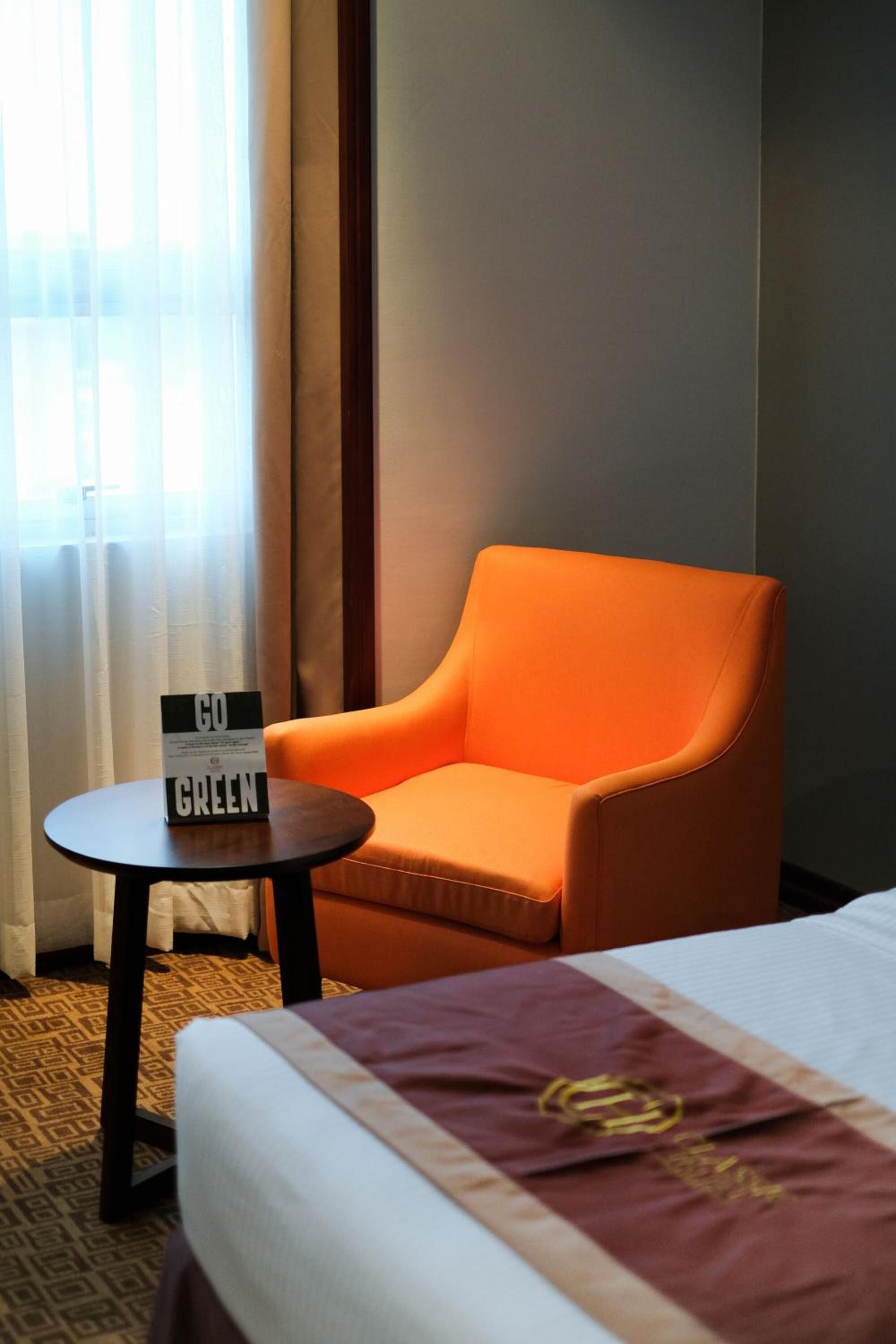 Una habitación de hotel con una cama, un sillón naranja, una mesita con un cartel y una ventana con cortinas.