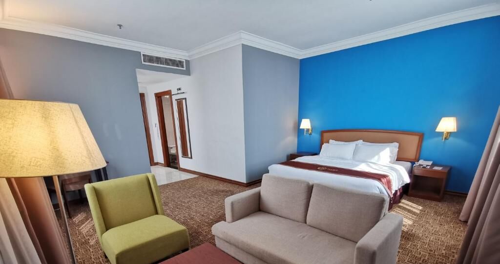 Una habitación de hotel con una cama, dos lámparas de noche, un sillón verde, un sofá color crema y una alfombra estampada.
