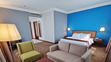Una habitación de hotel con una cama, dos lámparas de noche, un sillón verde, un sofá color crema y una alfombra estampada.