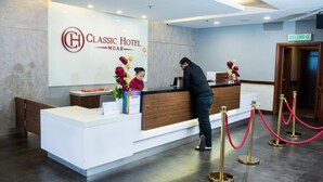 A hotel reception desk with a receptionist and a guest. The reception area has a modern design with a white counter, a wooden panel, and a sign that reads "Classic Hotel Muara." There are flower arrangements on the counter, and a sign indicating the direction to the "KELUAR" room. - Classic Hotel Muar (Muar)