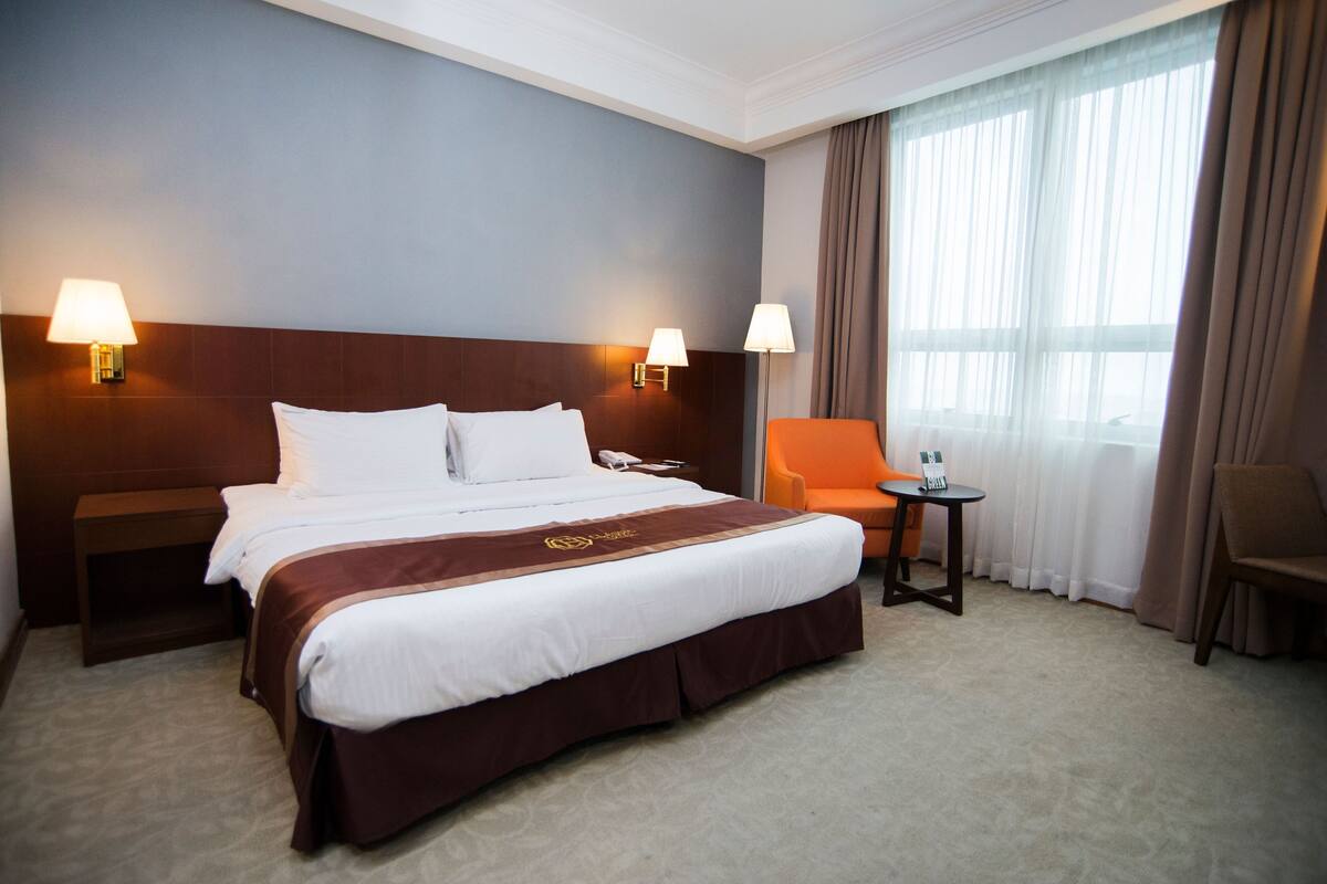 A hotel room with a large bed, two bedside lamps, a small table, a chair, and a window with curtains.