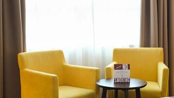 Camera d'albergo con due poltrone gialle, un tavolino rotondo e un cartello 'No Smoking', e un letto con testata bianca.