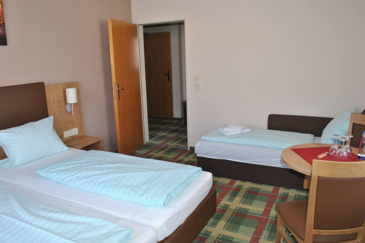 A hotel room with two beds, a wooden nightstand with a lamp, a plaid-patterned carpet, and a small table with a red placemat.
