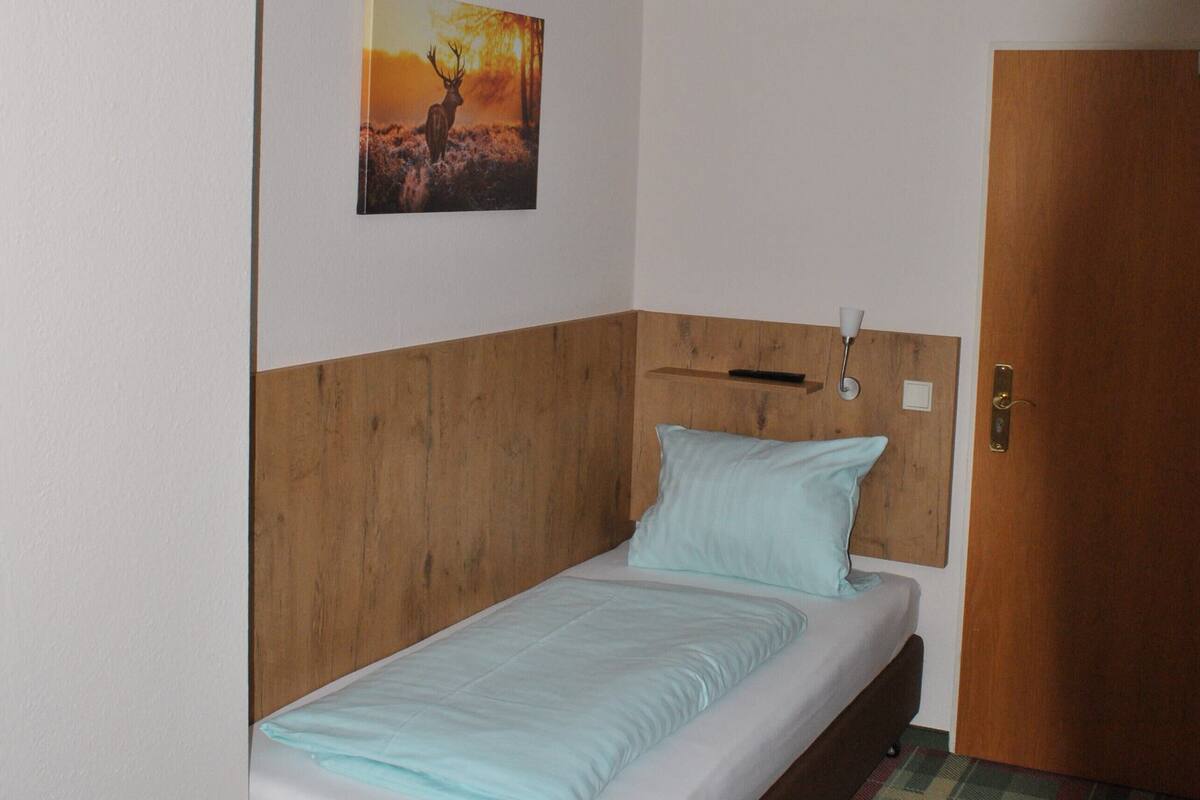 A single bed with a wooden headboard, a plaid carpet, a wall-mounted picture of a deer, and a door to the right.