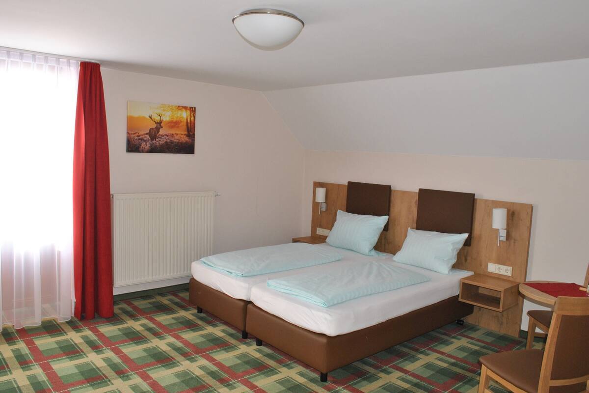 A double bed with two pillows, a nightstand, a chair, a red curtain, a radiator, and a painting of a deer.