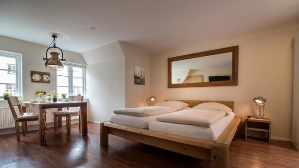 A spacious bedroom with a large bed, wooden floor, a dining area with a table and chairs, and a large window providing natural light. - Kajüte Am Nordermarkt (Flensburg)