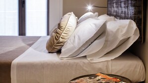 Een net opgemaakt bed met wit en beige beddengoed, een tijdschrift op een nachtkastje, en een raam met natuurlijk licht op de achtergrond.