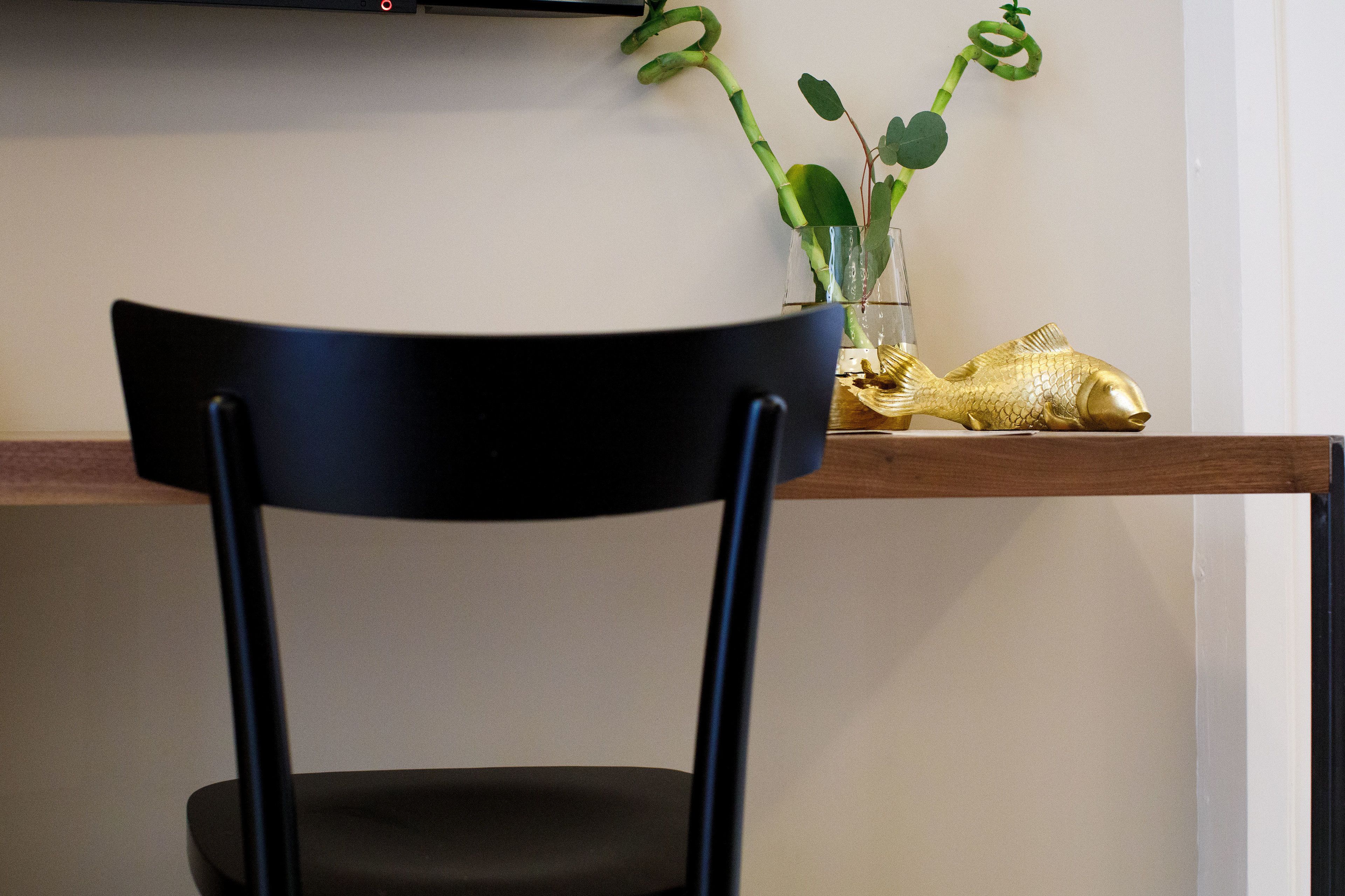 Une chaise noire est placée au premier plan, avec une étagère en bois derrière. Sur l’étagère, on aperçoit un vase en verre transparent contenant une plante verte et une décoration en forme de poisson doré.