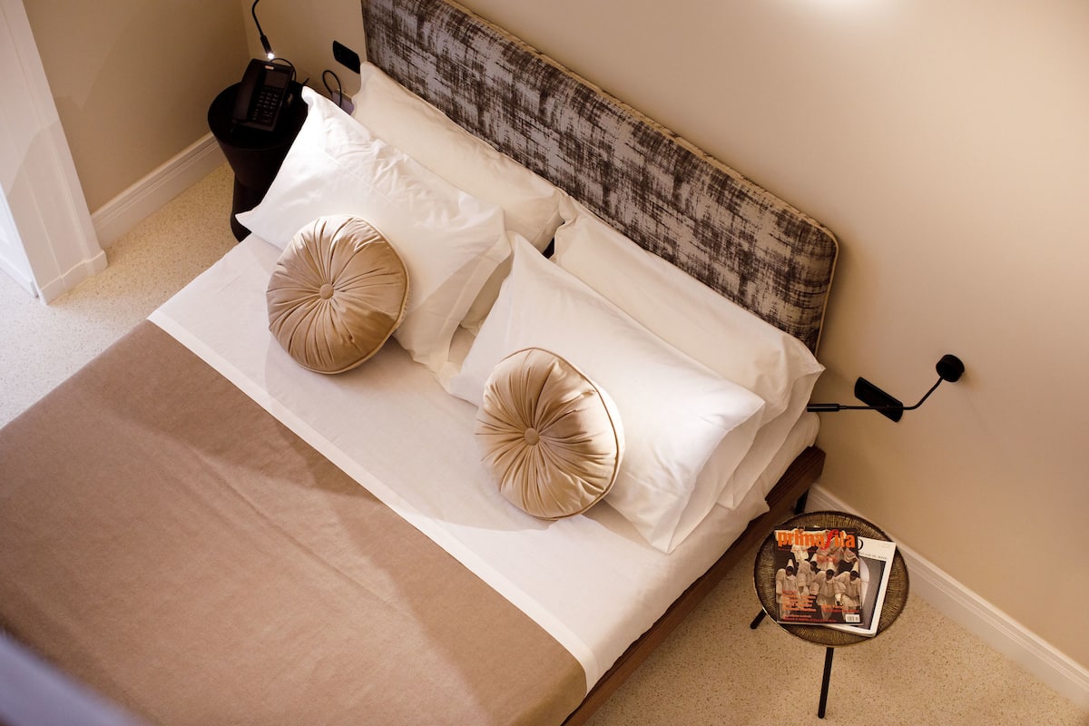 Una cama bien hecha con sábanas blancas y dos almohadas color beige, una mesita de noche con una revista, y una mesita auxiliar de pared con un teléfono.