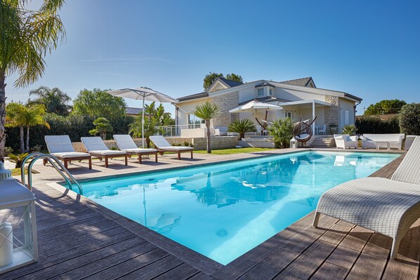 Piscina com espreguiçadeiras, um deck de madeira e uma casa ao fundo.