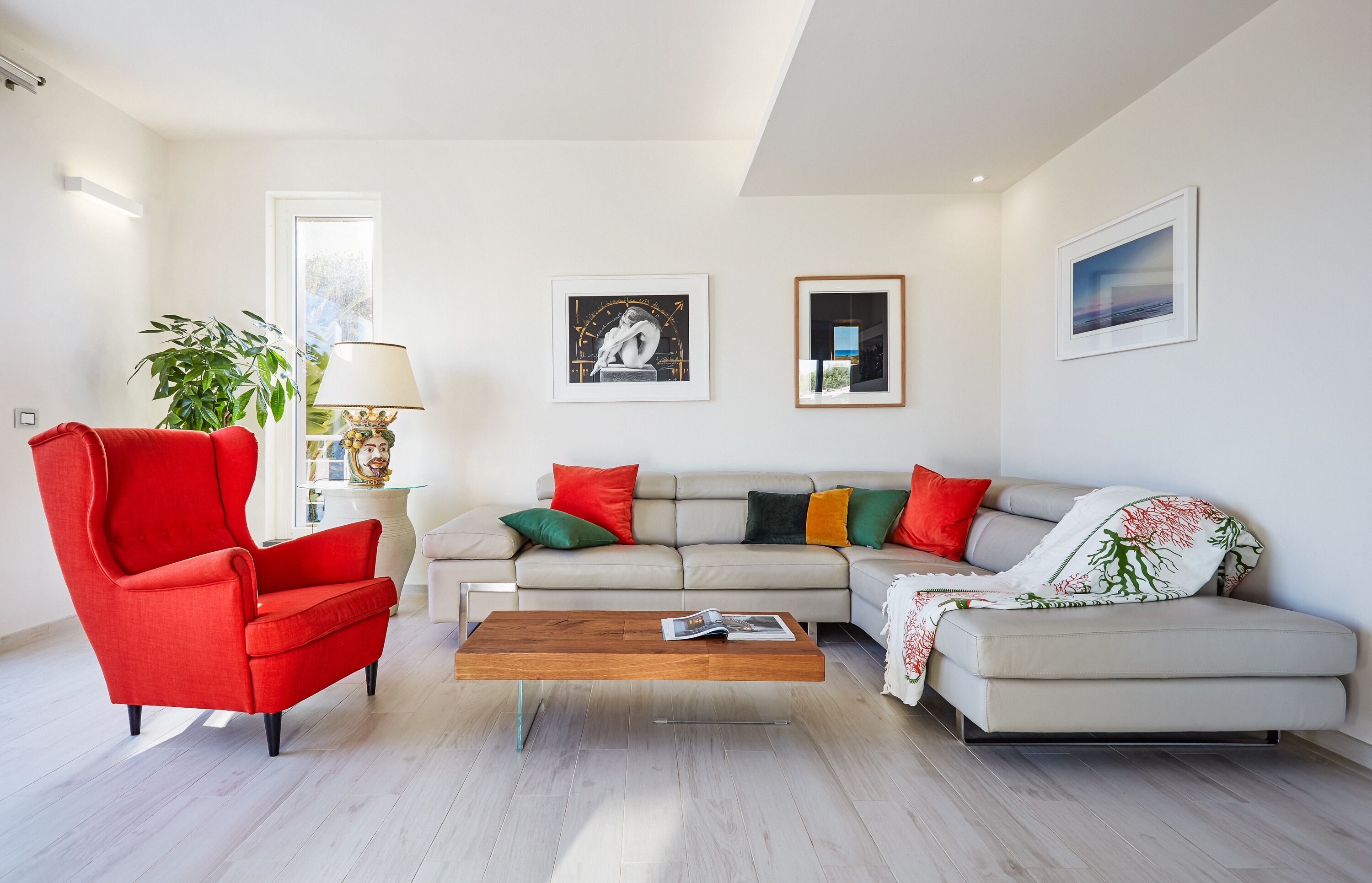 Een moderne woonkamer met een rode fauteuil, een beige bank met kleurrijke kussens, een houten salontafel en twee kunstwerken in lijsten aan de muur.