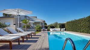 Zona de piscina con sillones de descanso y sombrillas, una terraza de madera y una piscina azul cristalina.