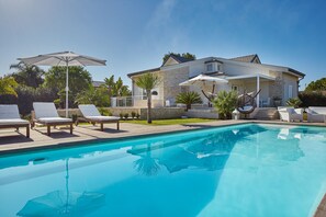 Uma piscina com espreguiçadeiras e guarda-sóis, uma casa de pedra com varanda, e um céu azul límpido.