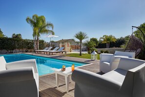 Área à beira da piscina com deck de madeira, espreguiçadeiras brancas e uma mesa com bebidas.