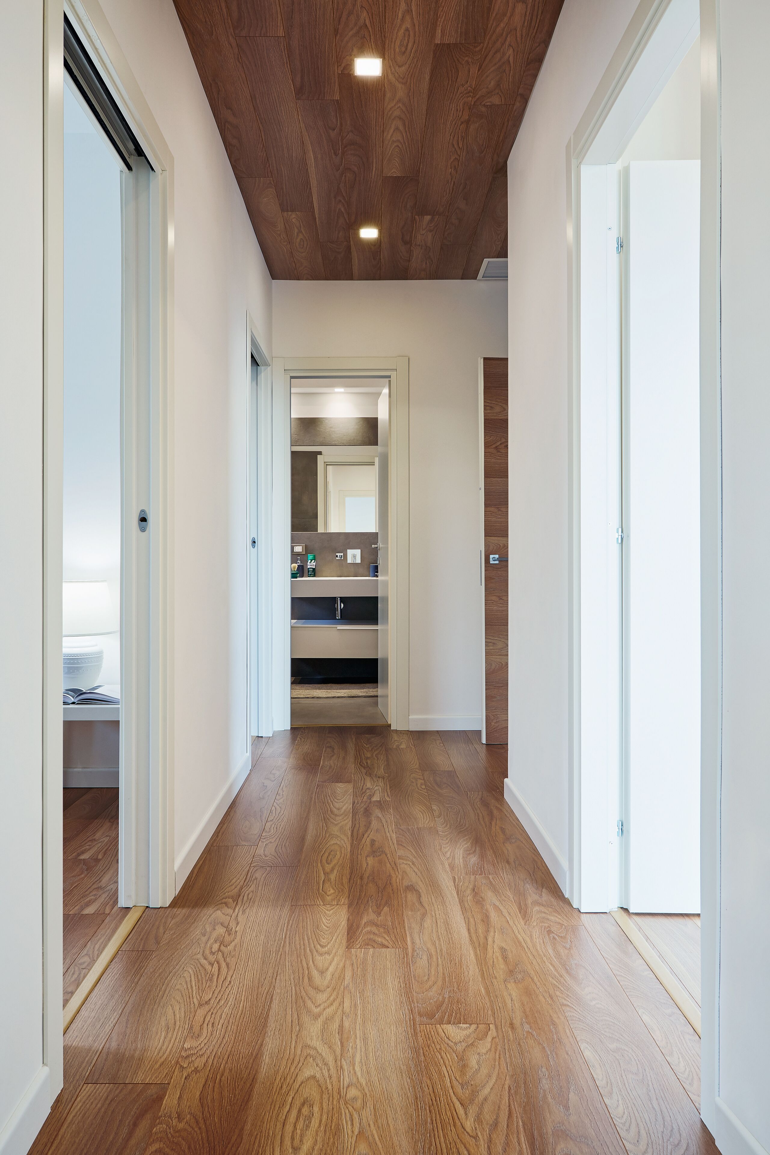 A hallway with wooden flooring and white walls, leading to a kitchen area with a sink and cabinets.