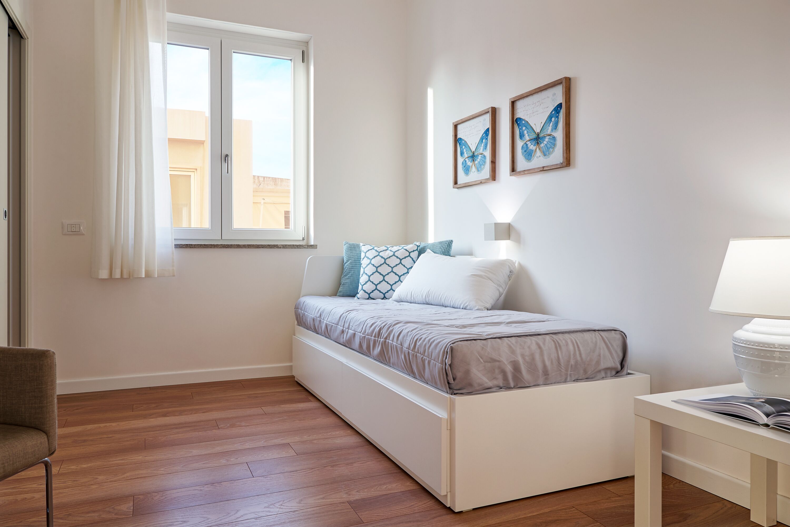 A bedroom with a bed, a bedside table, a chair, a window, and two framed butterfly pictures on the wall.
