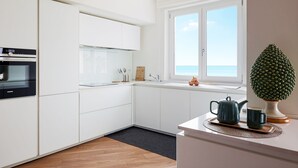 Une cuisine moderne avec des meubles blancs, un four encastré et une fenêtre donnant sur la mer.