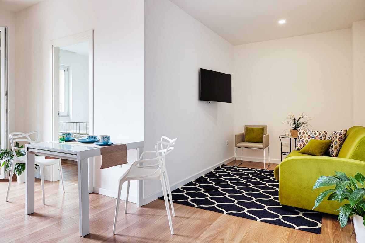 Un salon moderne comprenant un coin repas, un téléviseur à écran plat fixé au mur, un canapé vert et un tapis à motifs.