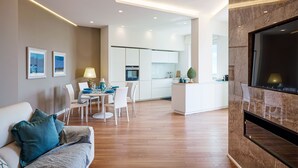 Un salon moderne avec un canapé, un coin repas comprenant des chaises et une table, une cuisine avec des meubles blancs et un téléviseur fixé sur un mur en marbre.