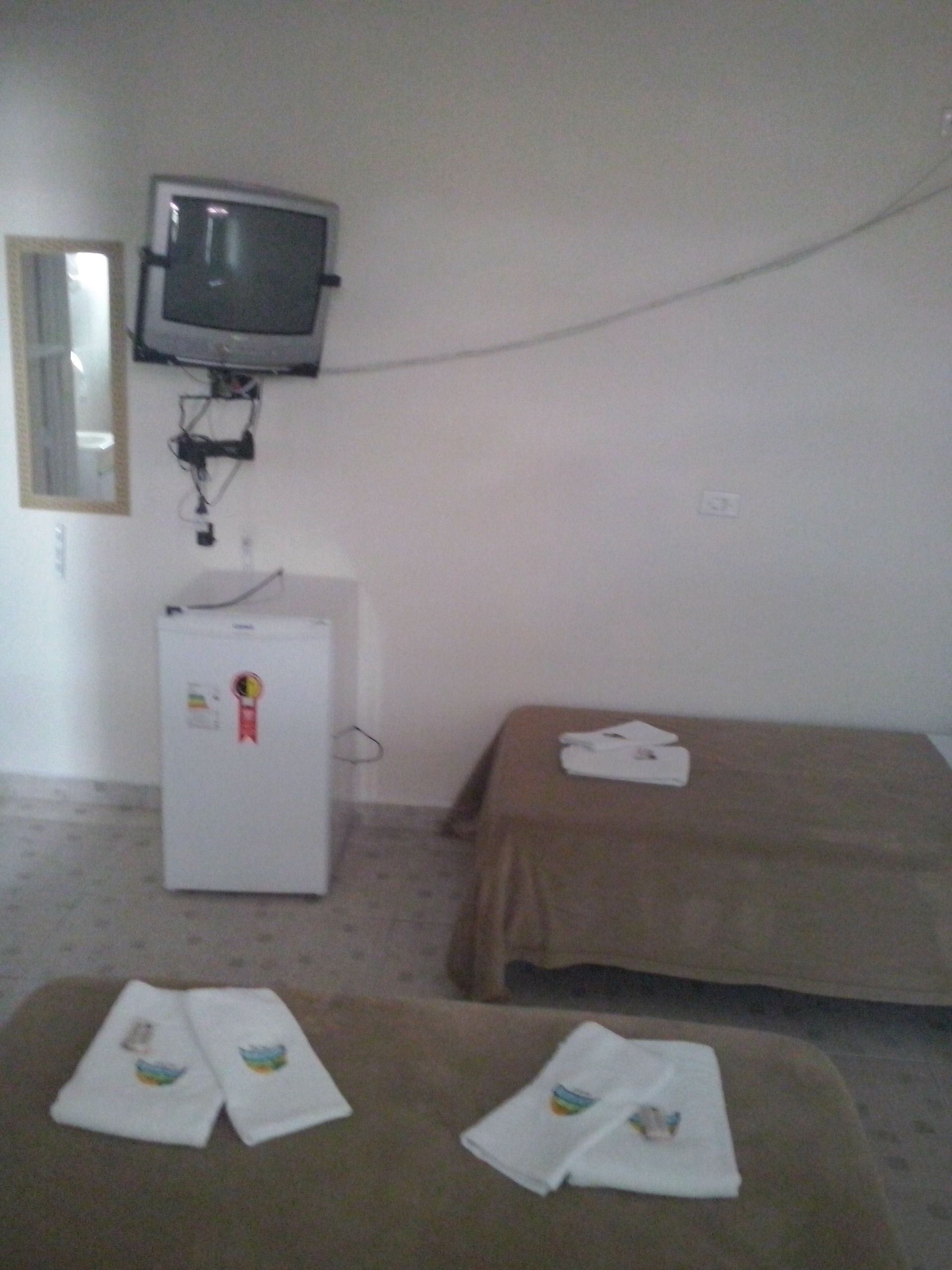 Una habitación pequeña con una cama, televisor montado en la pared, refrigerador y una mesa con toallas.