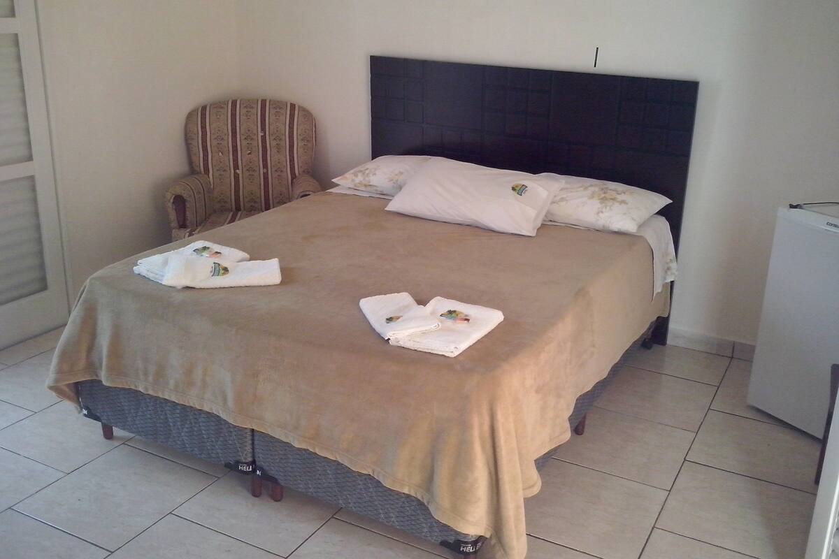 Una cama bien hecha con cabecera, una silla y un pequeño electrodoméstico blanco, probablemente un refrigerador, en una habitación con suelo de baldosas.